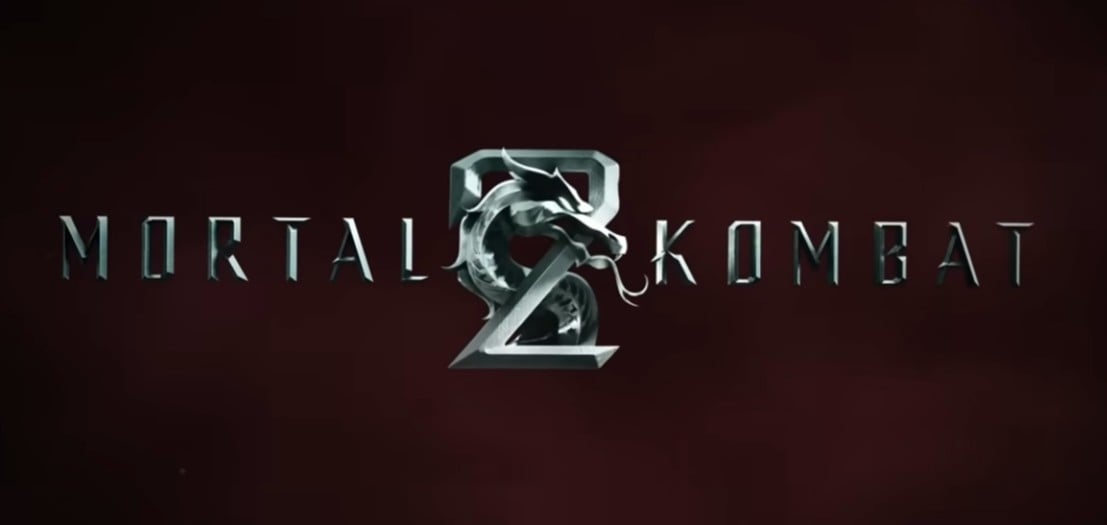 Logotipo oficial do filme Mortal Kombat 2 de 2026, com o título em letras prateadas texturizadas e o icônico dragão metálico no centro. O fundo é uma superfície texturizada vermelha escura e profunda.